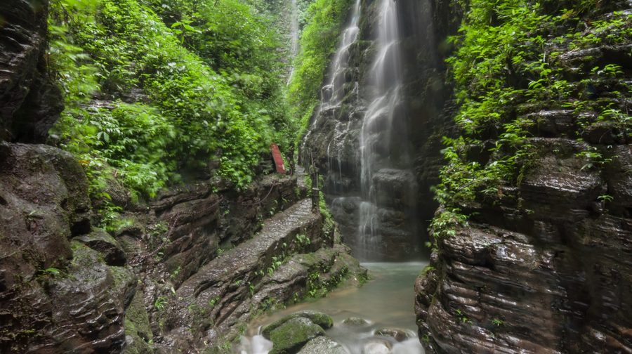 Sitting Dragon Gorge waterfall flowing through a lush green canyon with rocky cliffs and a narrow stone path.