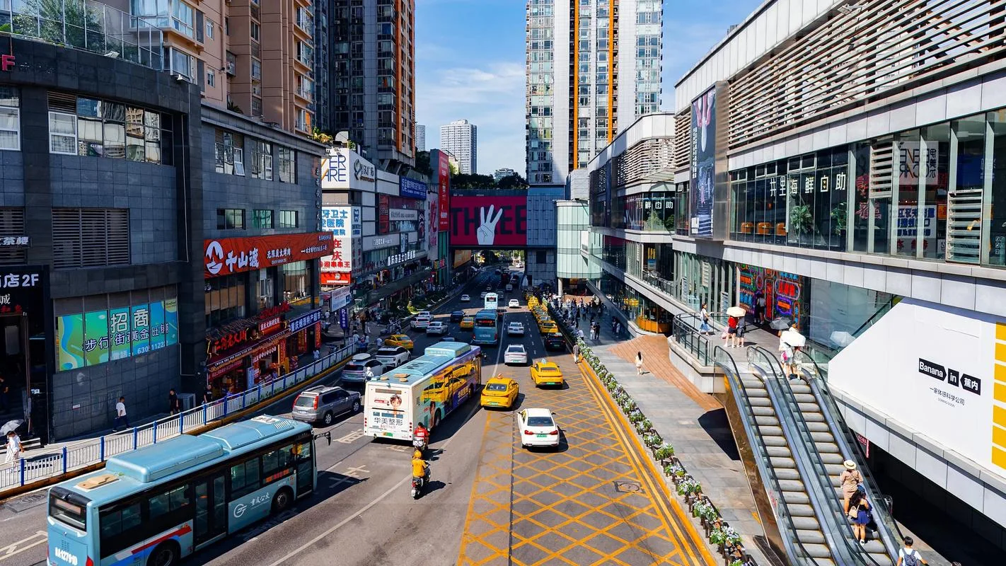 Busy Chongqing shopping street under strong midday summer sun, with buses and taxis passing